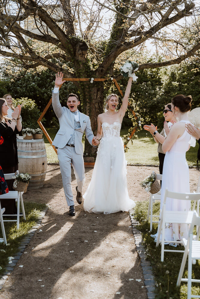 Brautpaar lacht und reißt die arme hoch beim Auszug seiner freien Trauung auf einem Weingut in der Pfalz
